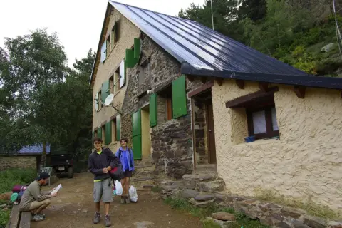 Llegada al refugio de Vallferrera. Dos jóvenes con mochilas y un hombre sentado revisando un mapa esperan en la entrada de piedra y estuco.