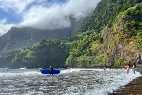 Surfer z niebieską deską na czarnej plaży Madera, w tle zielone klify i chmury.