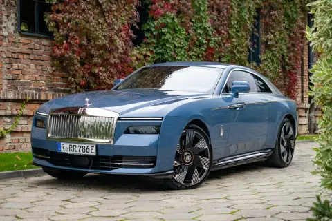Niebieski Rolls-Royce Spectre, luksusowy samochód elektryczny, zaparkowany przed ceglanym budynkiem porośniętym jesiennymi liśćmi.