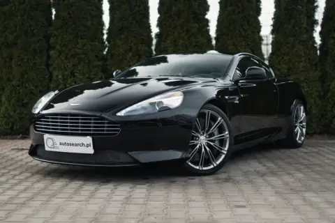 Czarny Aston Martin DB9 lśni na tle zielonych żywotników. Elegancki, sportowy samochód z charakterystycznym grillem i felgami.