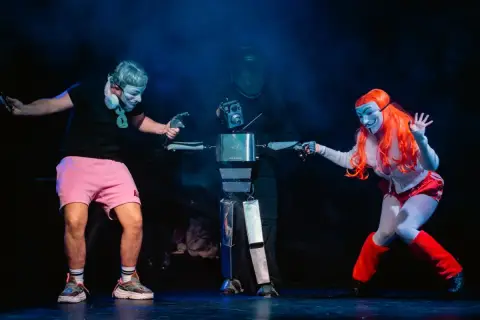Na scenie Opolskiego Teatru Lalki i Aktora trzej artyści w maskach, jeden z nich steruje robotem.
