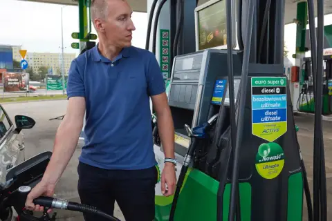 Mężczyzna tankuje samochód, pokazując, jak zatankować auto. Widoczna jest dystrybutor paliwa z różnymi rodzajami paliwa.