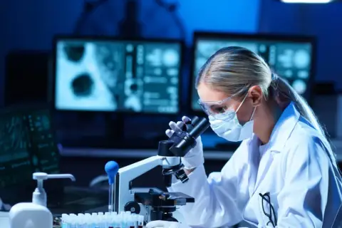 Diagnosta laboratoryjny w okularach i masce bada próbki pod mikroskopem. Na ekranach komputera widać wyniki badań.