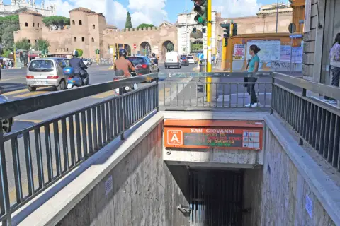 Ingresso metro San Giovanni, collegamento metro A. Traffico intenso con auto e scooter, sullo sfondo le antiche mura romane.