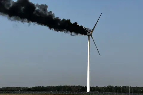 Płonąca turbina wiatrowa z gęstym, czarnym dymem unoszącym się na tle błękitnego nieba.