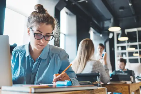Kobieta w okularach pracuje przy laptopie, pisząc w notatniku. W tle inni pracują, tworząc atmosferę skupienia od dziewiątej do piątej.