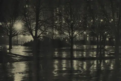 Nocny park, mokre alejki odbijające światła latarni i okien. Piktorializm w fotografii oddaje nastrój i atmosferę.