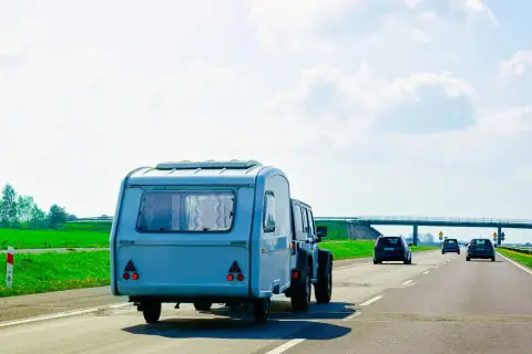 Samochód terenowy ciągnie dmc przyczepy kempingowej po autostradzie. W tle zielone pola i most.