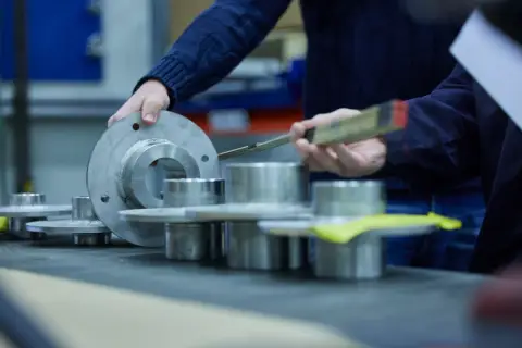Hands examine metal components, possibly for a fluid power equation application, with a ruler for measurement.