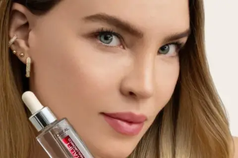 Mujer con cabello rubio y joyas brillantes sostiene un frasco de sérum, parte de su rutina de cuidado de la piel.