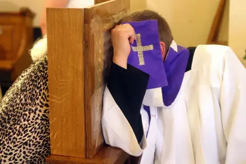 Spowiedź w konfesjonale. Czy nie pójście do kościoła z powodu choroby to grzech? Kapłan w białej albie i fioletowej stuły z krzyżem.