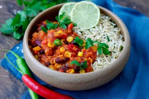 Herzhaftes Chili sin Carne mit Reis, garniert mit frischer Petersilie und Limettenspalten. Rote und grüne Chilis liegen daneben.