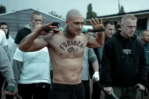 Wytatuowany mężczyzna z napisem "Furioza" na klatce piersiowej, otoczony przez grupę mężczyzn, przygotowuje się do konfrontacji.