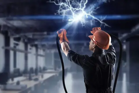 Elektryk w kasku i rękawicach trzyma przewody, z których wydobywa się potężne przepięcie elektryczne.
