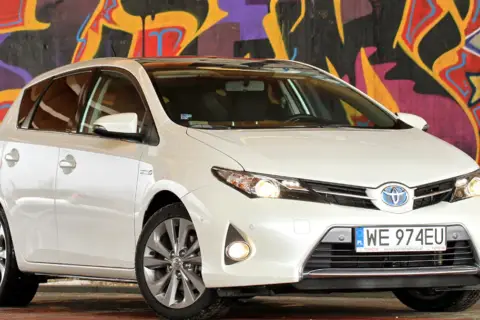 Biała Toyota Auris 2015 z polskimi tablicami rejestracyjnymi WE 974EU zaparkowana przed kolorowym muralem.