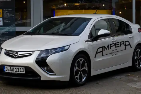Biała Opel Ampera z czarnym napisem "AMPERA" na drzwiach, zaparkowana przed salonem samochodowym.