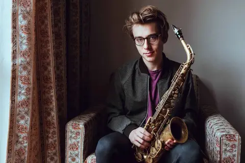 Młody muzyk z saksofonem, przyszłość najwybitniejszych polskich muzyków jazzowych, siedzi w fotelu.