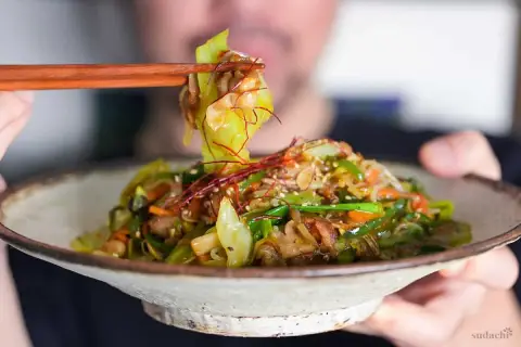 A person uses chopsticks to lift a bite of a colorful Japanese stir fry, featuring cabbage, carrots, and peppers, from a rustic bowl.
