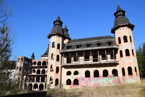 Niedokończony zamek w Łapalicach, z ceglanymi wieżami i łukowymi oknami, pokryty graffiti, pod błękitnym niebem.