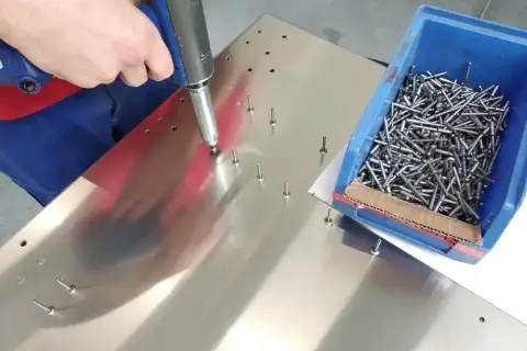 Ręka pracownika używa narzędzia do montażu nitów zrywalnych na metalowej powierzchni. Obok znajduje się niebieskie pudełko pełne nitów.