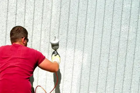 Uomo con maglietta rossa e occhiali da sole sta pitturando casa a spruzzo con una pistola.
