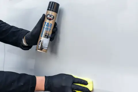Ręce w czarnych rękawiczkach używają preparatu K2 Tar Remover, by usunąć klej z szyby auta.