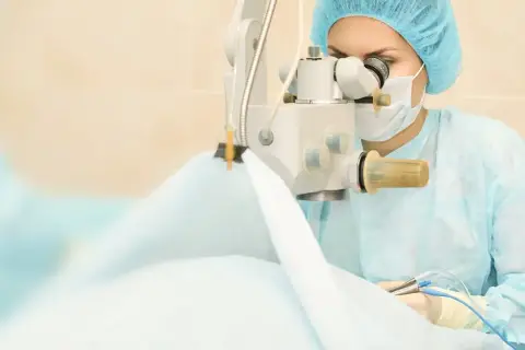Chirurg w masce i czepku wykonuje operację zaćmy, używając mikroskopu i precyzyjnych narzędzi.
