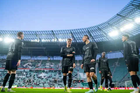Piłkarze w czarnych strojach na murawie stadionu. Jeden z nich pyta: "walkower co to?", ale koledzy skupiają się na grze.