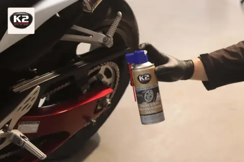 Ręka w czarnej rękawiczce trzyma puszkę smaru do łańcucha K2. Motocykl w tle.