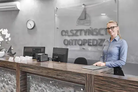 Recepcjonistka w okularach stoi przy ladzie w nowoczesnej klinice ortopedycznej.