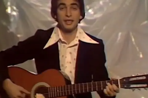 Mężczyzna w garniturze i wzorzystej koszuli gra na gitarze. W tle widać materiał, na którym **ciągle pada tekst**.