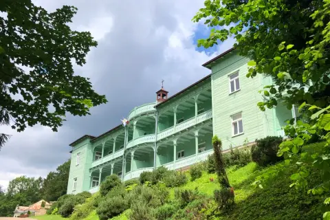 Zielony, zabytkowy klasztor w Komańczy, otoczony bujną zielenią, z charakterystycznymi werandami i białymi balustradami.