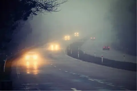 Samochody na mglistej drodze, ich światła przeciwmgielne przebijają się przez gęstą mgłę, tworząc świetlistą poświatę.