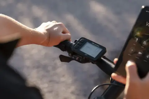 Rowerzysta trzyma kierownicę, na której zamontowany jest licznik rowerowy nie pokazuje prędkości. Obok wyświetlacz telefonu z połączeniem do Valerie.