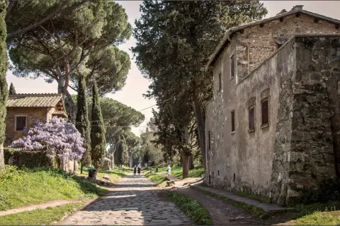 Une ancienne voie romaine à Rome, bordée de cyprès et de pins parasols, avec des maisons en pierre et des glycines en fleurs.