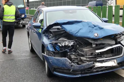 Niebieski samochód po kolizji drogowej, z przodu mocno zniszczony. Na drodze stoją ludzie w kamizelkach odblaskowych.