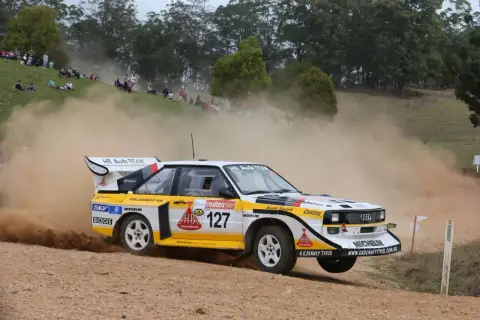 Klasyczny Audi Quattro z grupy B, oznaczony numerem 127, pędzi po szutrowej drodze, wzbijając tumany kurzu.