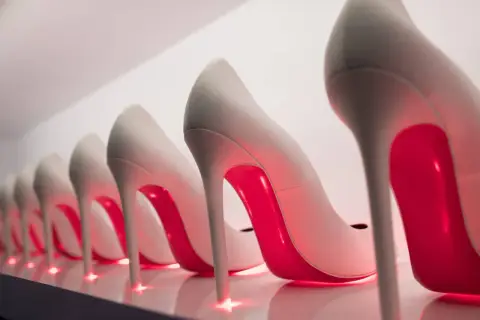 Rząd eleganckich, jasnych szpilek z czerwonymi podeszwami, przypominających kultowe Louboutiny, lśni w blasku neonowego światła.