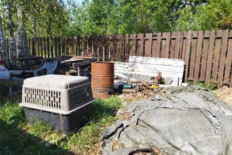 Zarośnięty ogród z rupieciami, beczką i klatką dla psa. Wygląda jak podwórko uciążliwego sąsiada na wsi.