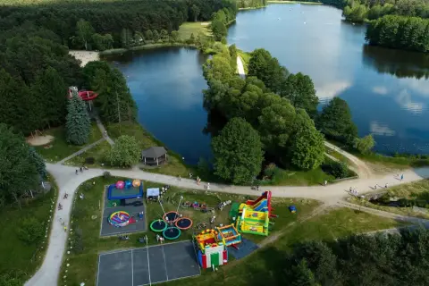 Rekreacja nad jeziorem w Bełchatowie: plac zabaw z dmuchańcami, trampolinami i zjeżdżalnią wodną, otoczony lasem i wodą.