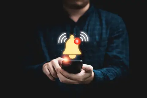 Dłoń trzymająca smartfon, nad którym unosi się złoty dzwonek z powiadomieniem.
