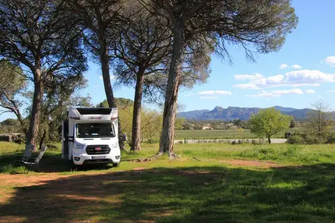 Un camping-car blanc est garé sous des pins, avec des chaises pliantes à côté. Un paysage de vignobles et de montagnes s'étend au loin. Un guide aire camping car idéal pour l'aventure.