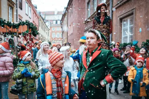Dawid w stroju elfa prowadzi świąteczny korowód ulicą pełną ludzi i ozdób. Dzieci z uśmiechem patrzą na magię jarmarku.