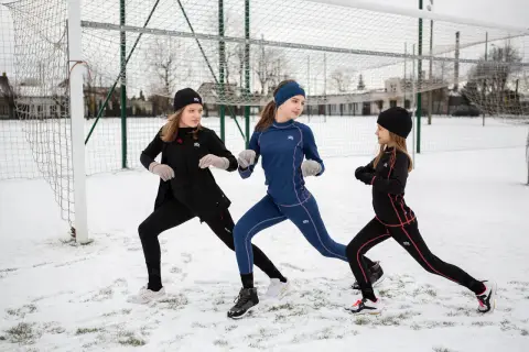 Trzy dziewczynki w czarnych i granatowych koszulkach termoaktywnych dziecięcych ćwiczą na śniegu przy boisku.