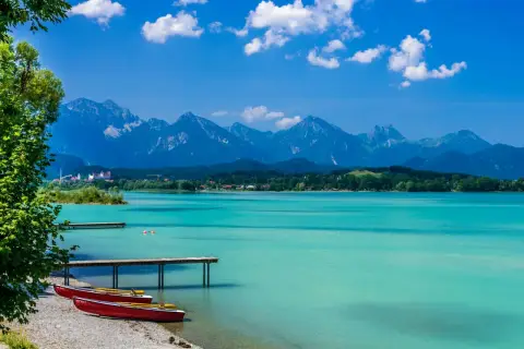 Türkisblaues Wasser am Badesee Bayern, mit roten Ruderbooten am Kiesstrand und majestätischen Alpen im Hintergrund.