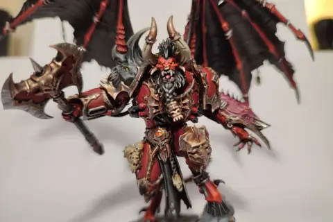 Groźny chaos daemon prince z czerwono-złotą zbroją, czarnymi skrzydłami i toporem.