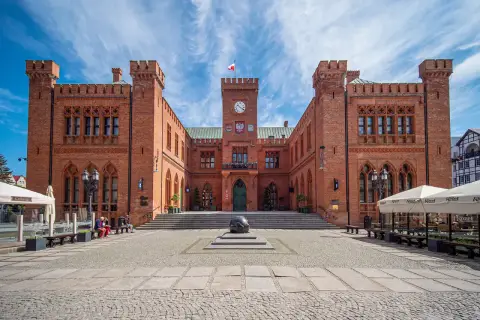 Rynek Kołobrzeg: ceglany ratusz z zegarem i flagą, przed nim kamienna rzeźba i brukowany plac.