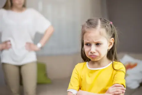 Smutna dziewczynka w żółtej bluzce, z matką w tle.