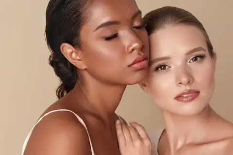 Dos mujeres, una con piel oscura y ojos cerrados, la otra con piel clara y ojos abiertos, comparten un momento íntimo sobre un fondo beige.