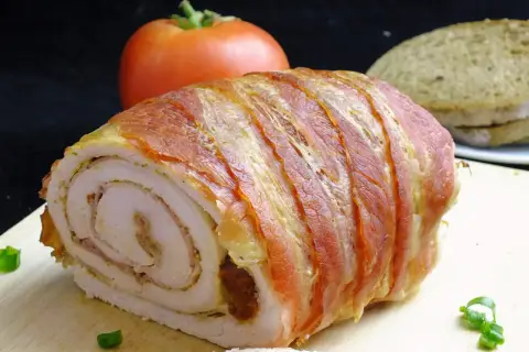 Rolada z indyka w boczku, idealna do krojenia bez rozpadania się. Na desce z boczku, obok pomidora i chleba.